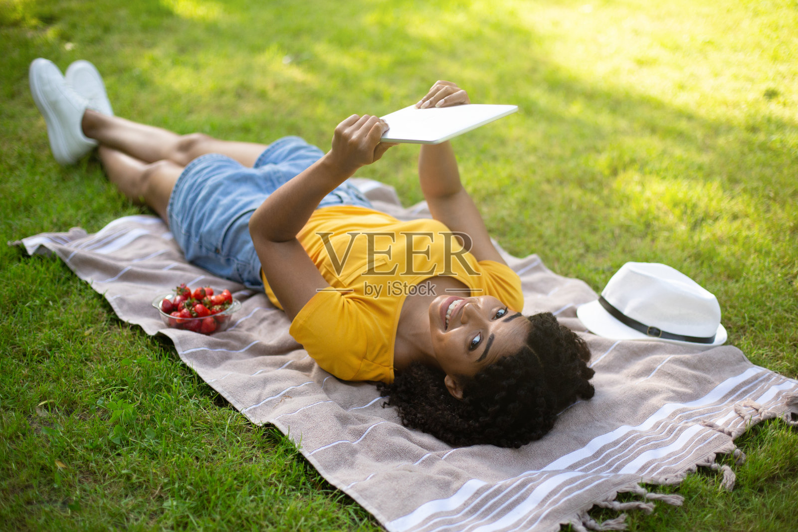 酷黑女孩躺在野餐毯子上,在外面的平板电脑上看视频照片摄影图片
