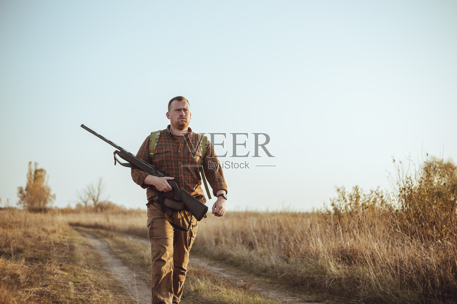 强壮的年轻猎人红胡子拿着他的枪,走在泥土路在蓝天下照片摄影图片