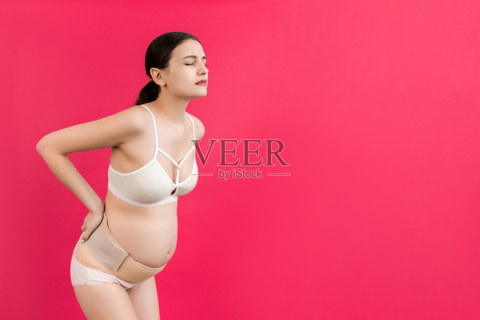 肖像的支持绷带对背痛的孕妇在粉红色的背景与复制空间。母亲背部疼痛。骨科腹部支撑带概念照片摄影图片