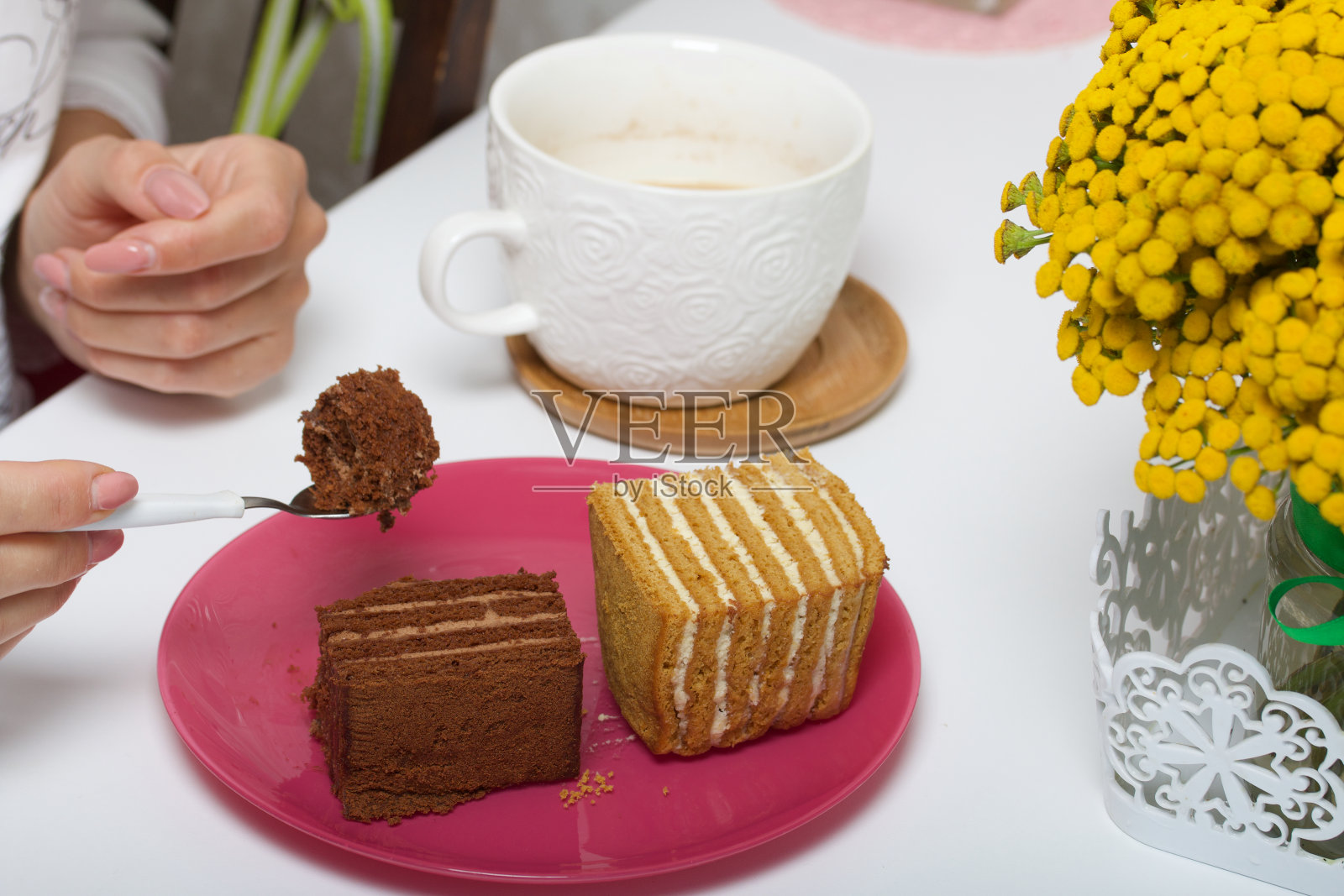 两片蜂蜜蛋糕。其中一个是用巧克力做的。一个女人早餐吃甜点。旁边是一杯牛奶茶和一束唐茜。照片摄影图片