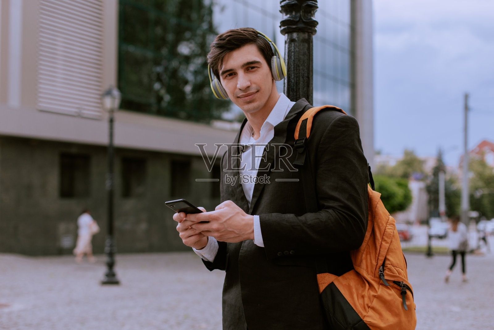 一名男子站在现代商业大厦前,背着背包,手里拿着智能手机,戴着耳机听着音乐照片摄影图片