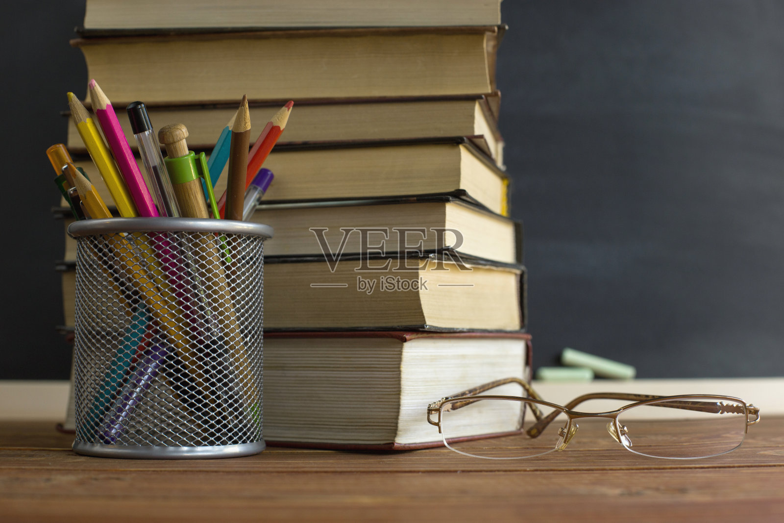 眼镜老师的书和一个架子上有铅笔的桌子,背景上有黑板和粉笔。教师节的概念。副本的空间。照片摄影图片