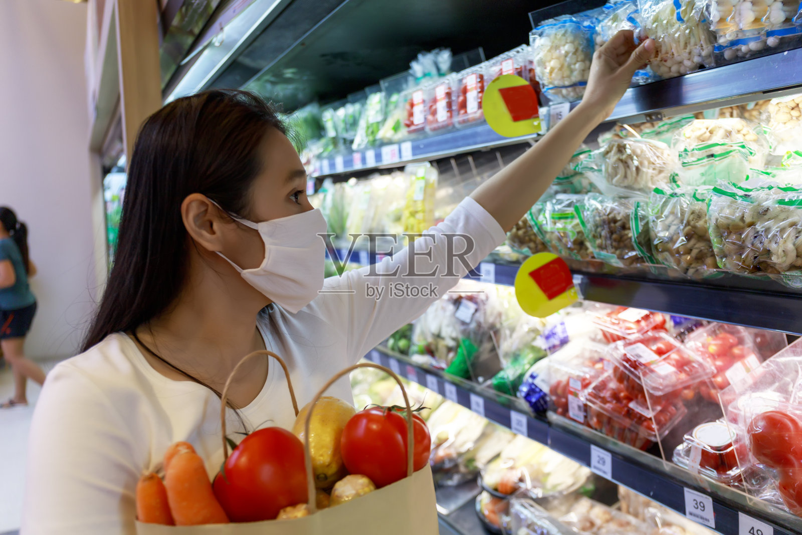 亚洲妇女戴着保护面罩,拿着装着水果和蔬菜的纸袋在超市购物。女孩,去杂货店买水果。新冠肺炎疫情后的新常态。家庭的概念。照片摄影图片