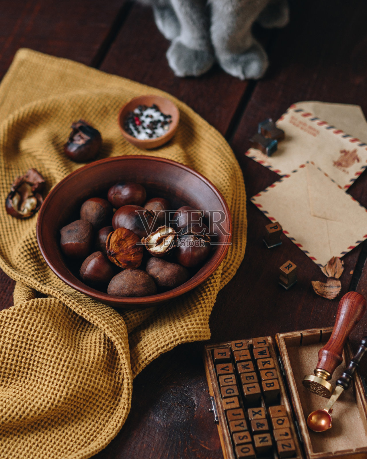 木桌上放着一碗烤栗子。有邮信封和邮票。背景中可以看到一只灰猫的腿。家里舒适。旅行。邮寄信件照片摄影图片