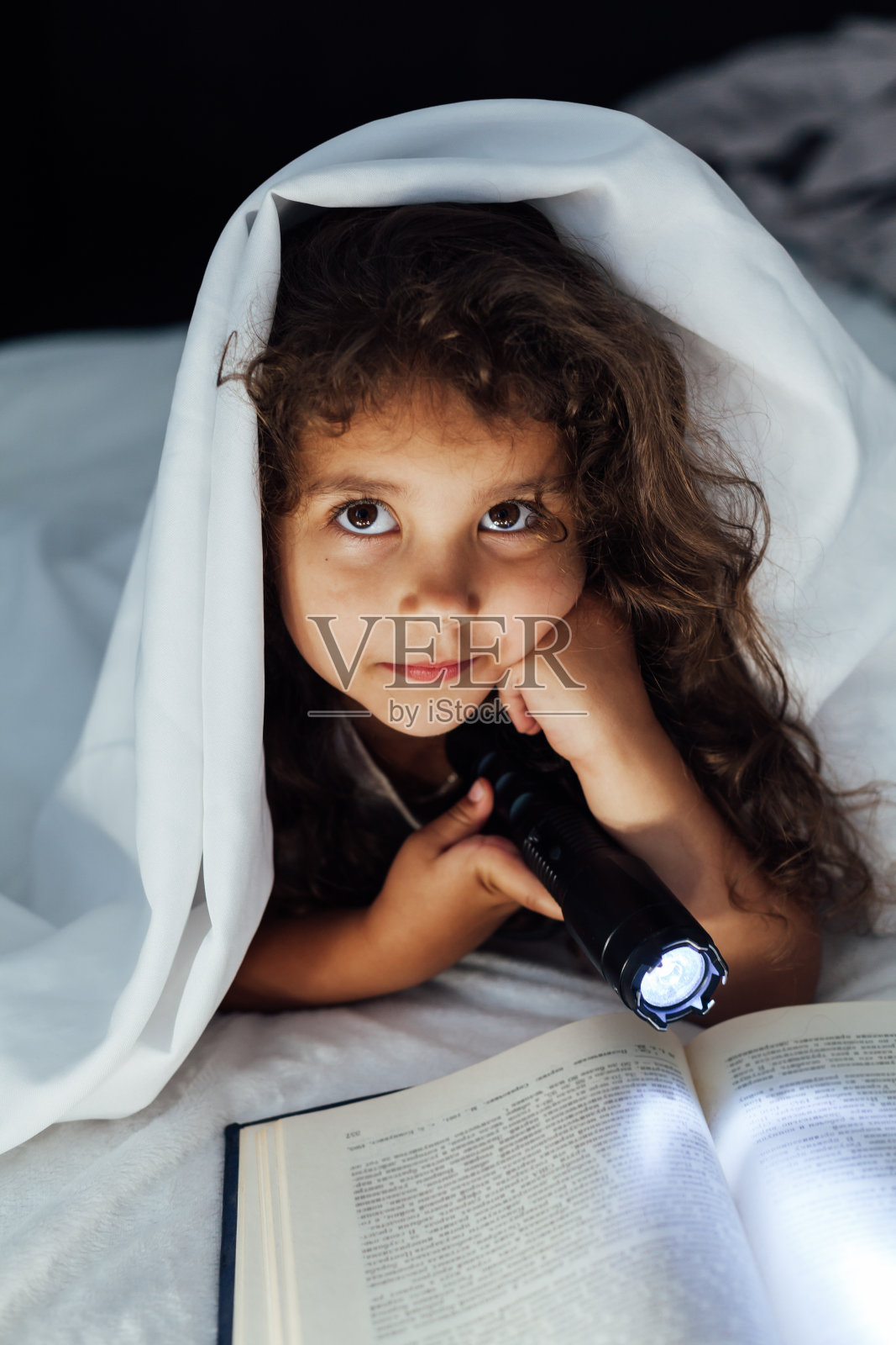 漂亮的小女孩在卧室的毯子下面拿着手电筒看书照片摄影图片