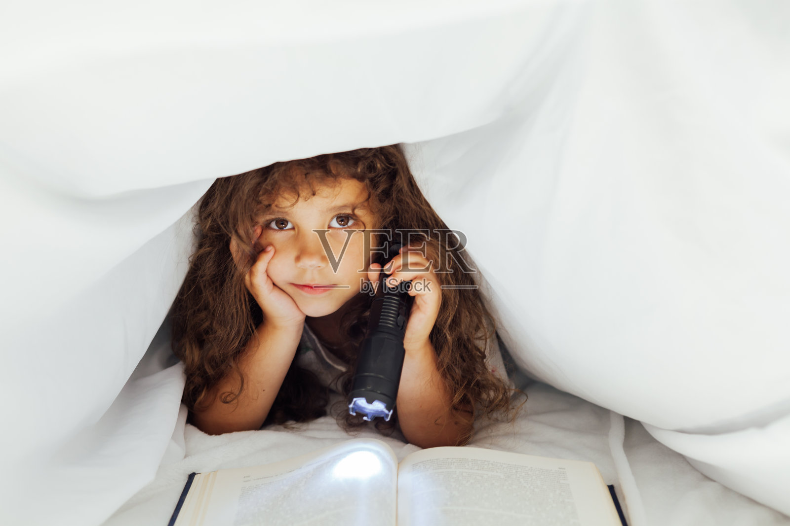漂亮的小女孩在卧室的毯子下面拿着手电筒看书照片摄影图片