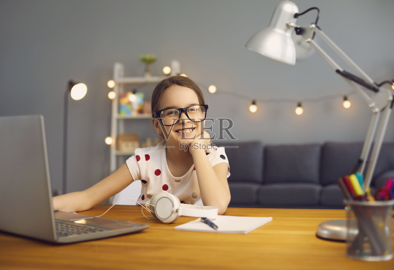 网上学校培训。戴着眼镜的聪明快乐的小女孩微笑着在房间里用笔记本电脑讲课。照片摄影图片
