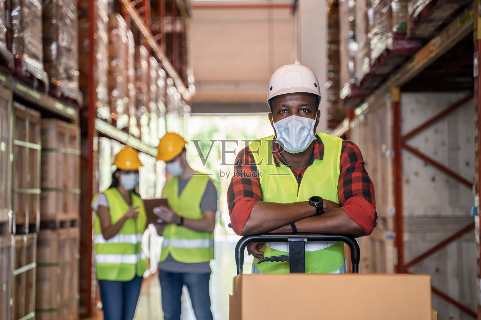 一群戴着防护面具在工厂仓库工作的工人。在新冠疫情危机期间,一名黑人推着金属车车,手里拿着盒子包裹。物流产业的概念。照片摄影图片