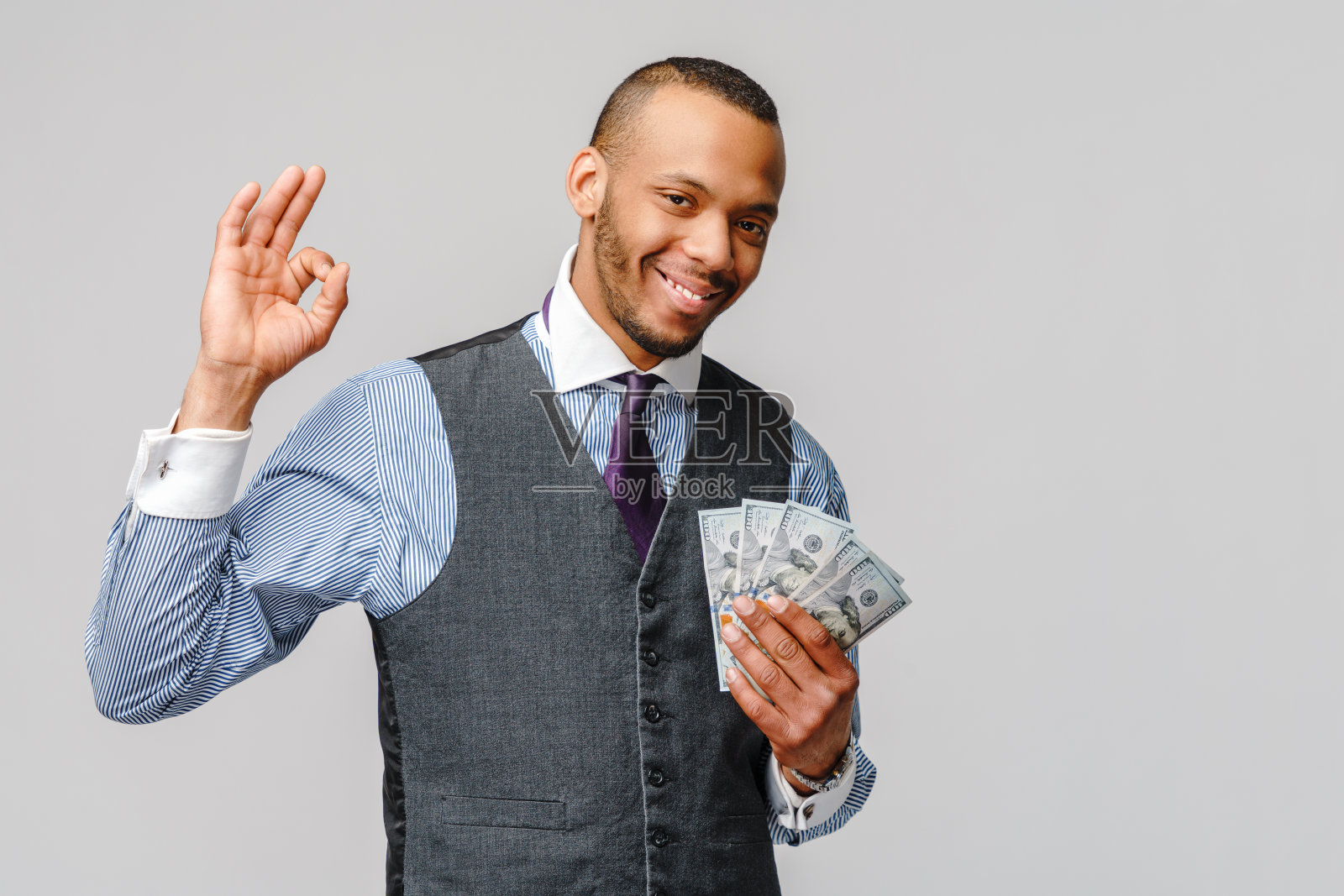 一个激动的年轻非洲裔美国人的肖像,手里拿着现金,在浅灰色的背景上显示OK的标志照片摄影图片