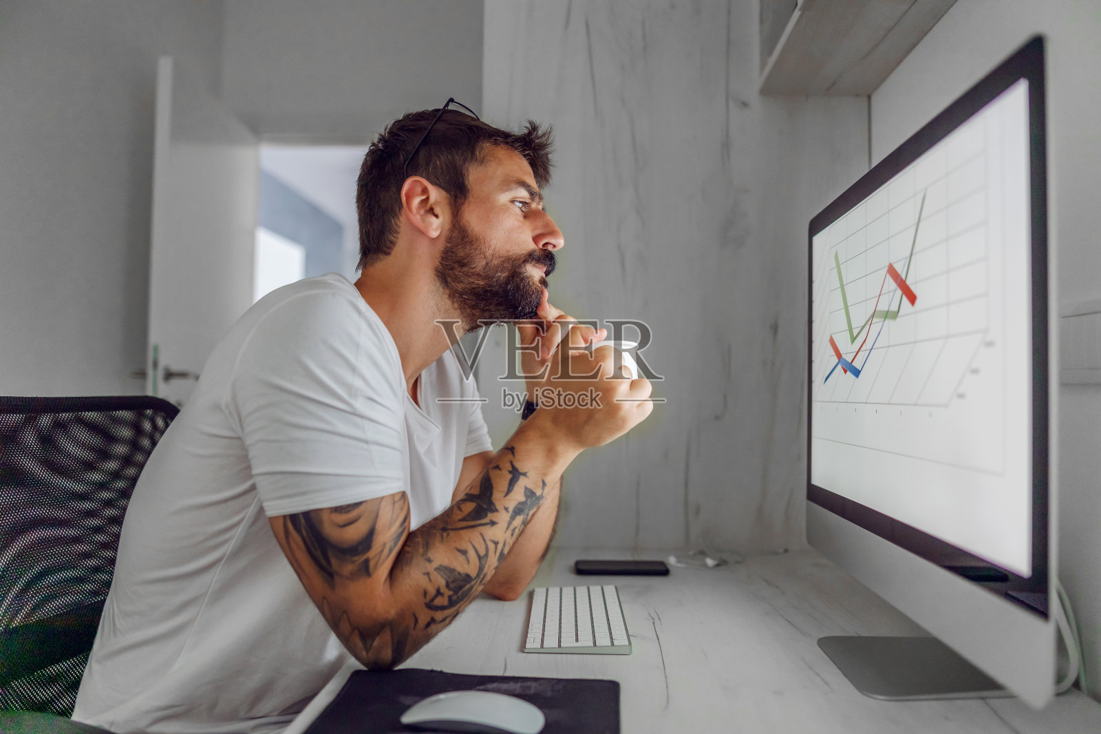 年轻英俊的留着胡子的员工坐在他的家庭办公室里分析股票市场的图表。冠状病毒爆发期间的隔离。照片摄影图片