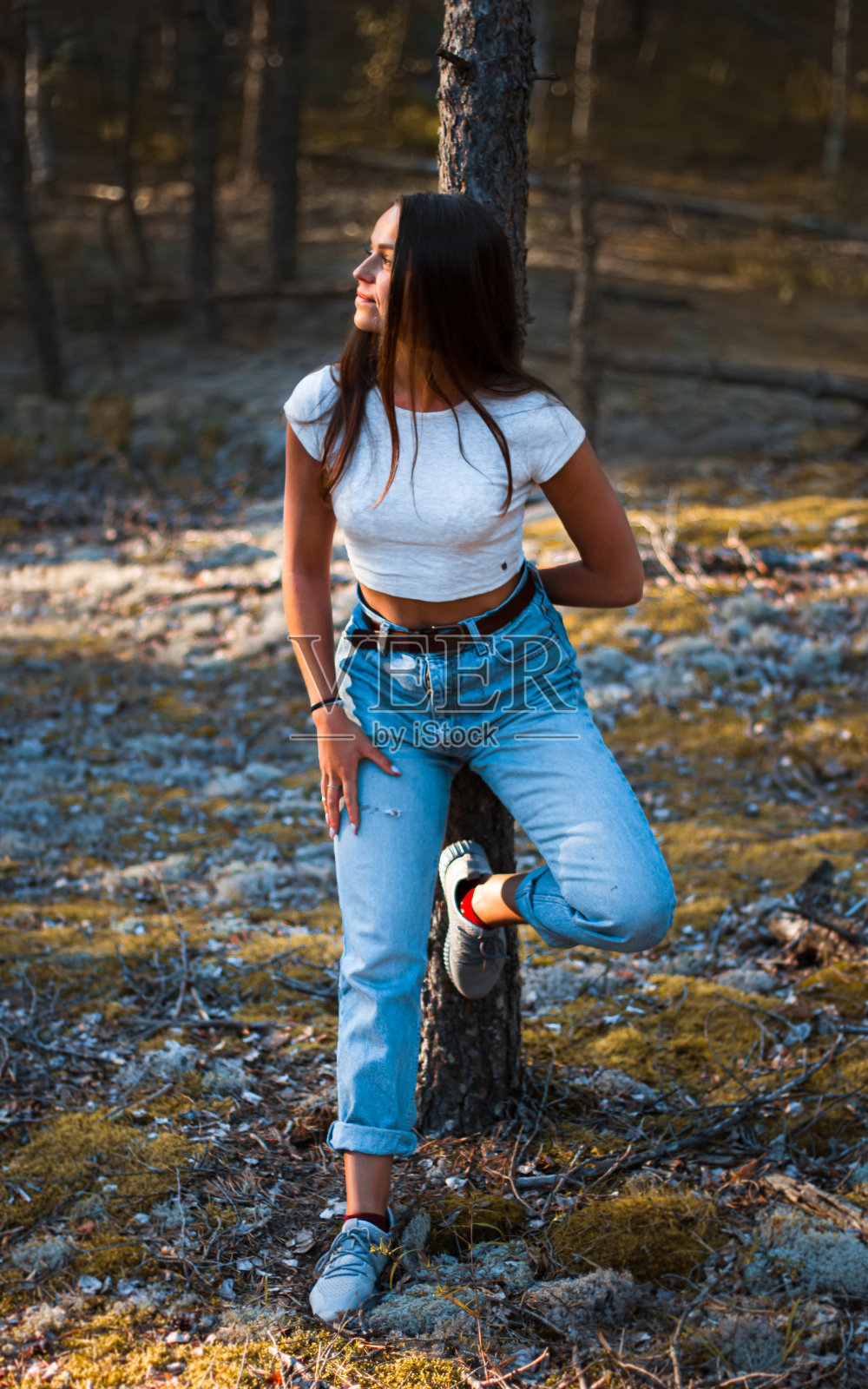 年轻时髦的漂亮女人在妈妈的牛仔裤和白色的顶部是倾斜和握住她的手在夏季松树林的一棵树照片摄影图片