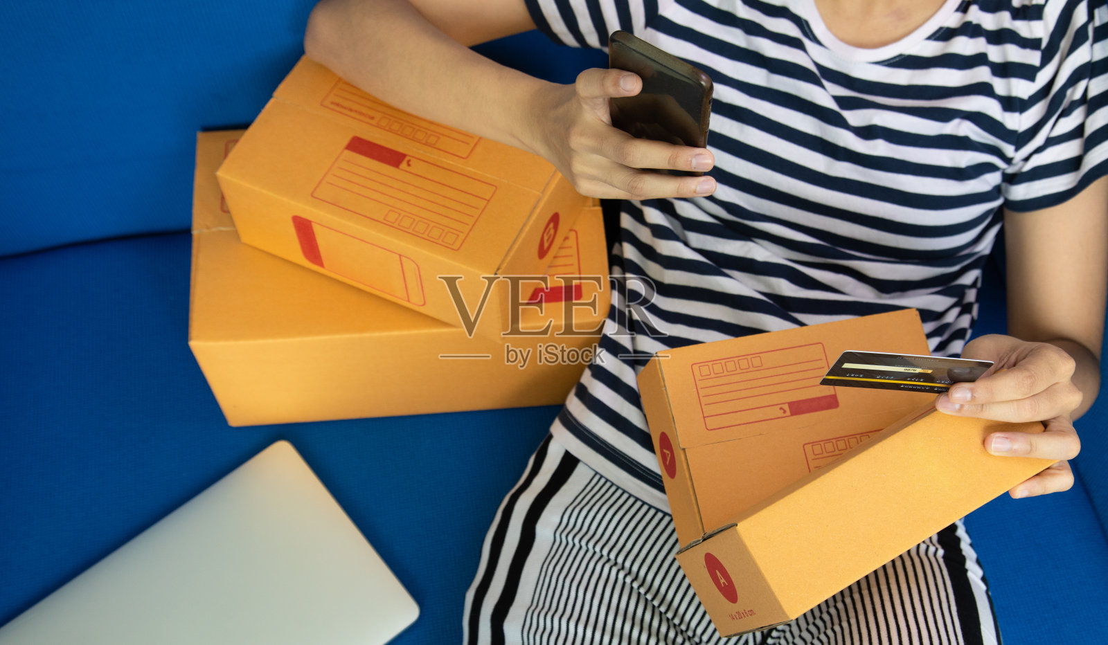 俯视图的十几岁的购物者是坐在沙发上,通过智能手机网上购物与信用卡的数字技术世界,看到她身边有很多盒子的衣服。照片摄影图片