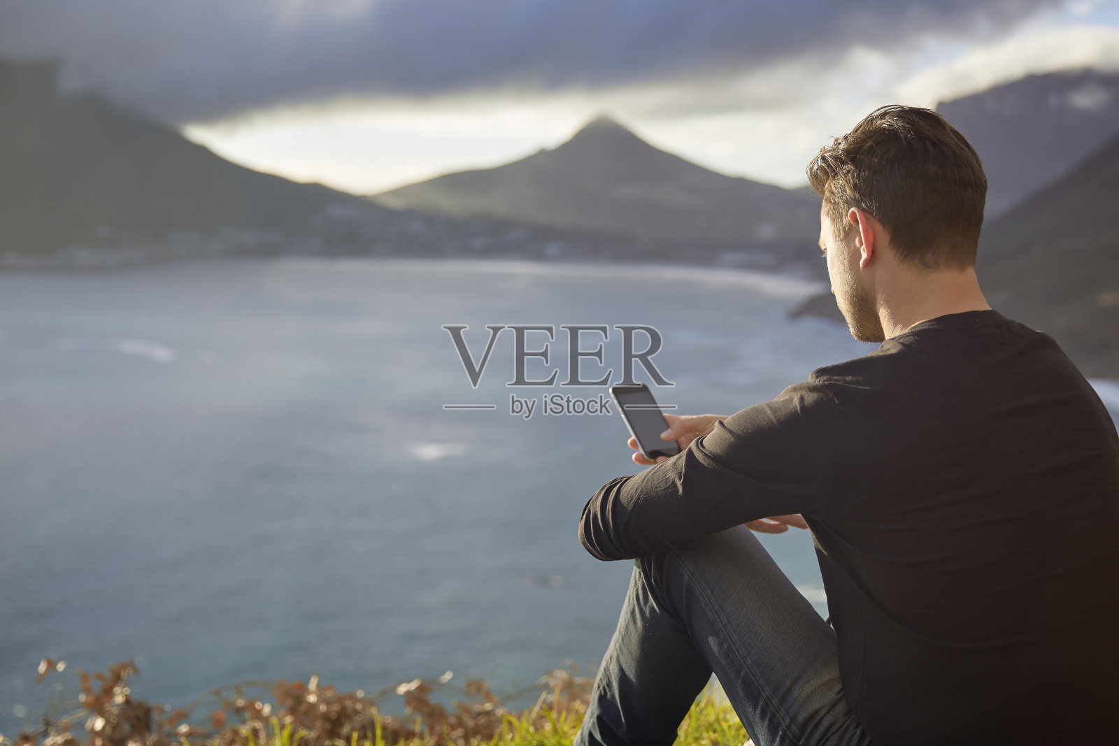 坐在悬崖边用智能手机的男人。照片摄影图片