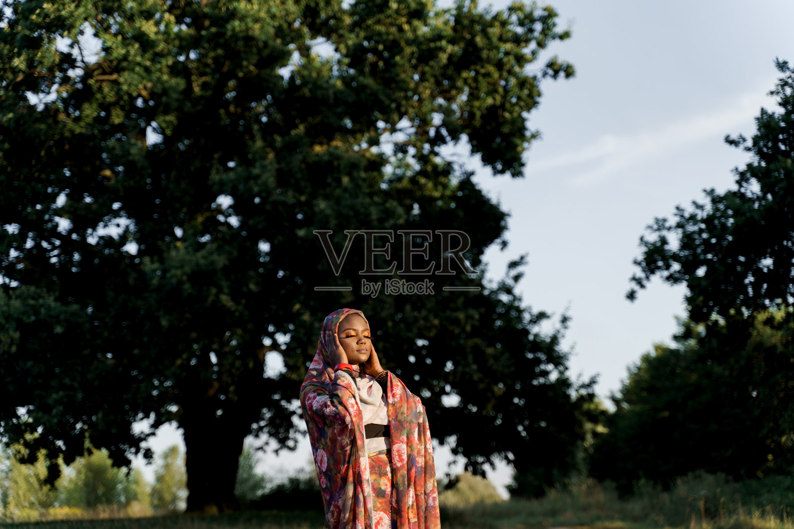 时尚迷人的穆斯林女孩在时尚的传统服装,妇女在传统的头巾与闭上眼睛触摸她的围巾。世界文化。非洲人在绿色草地上的生活方式。照片摄影图片