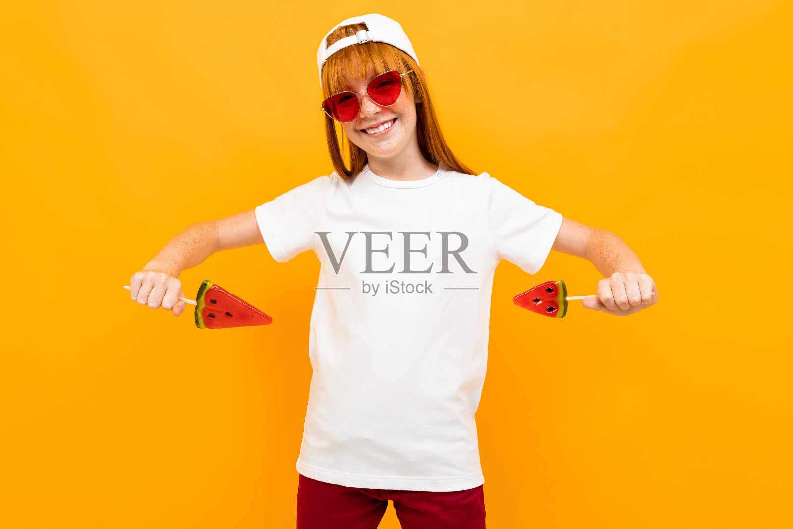 快乐的女孩红头发,白色的t恤,白色的帽子,微笑的西瓜棒棒糖孤立在黄色的背景照片摄影图片