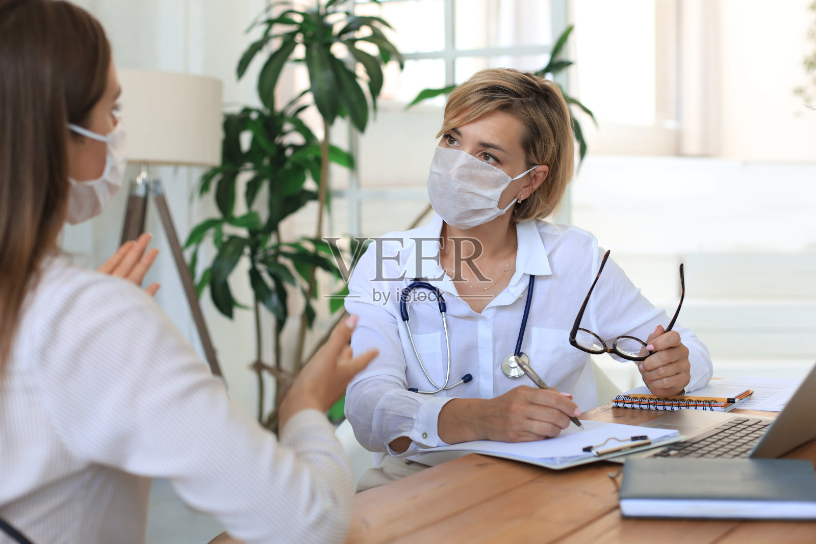 中年女性医师在医疗口罩上与病人在办公室咨询。照片摄影图片