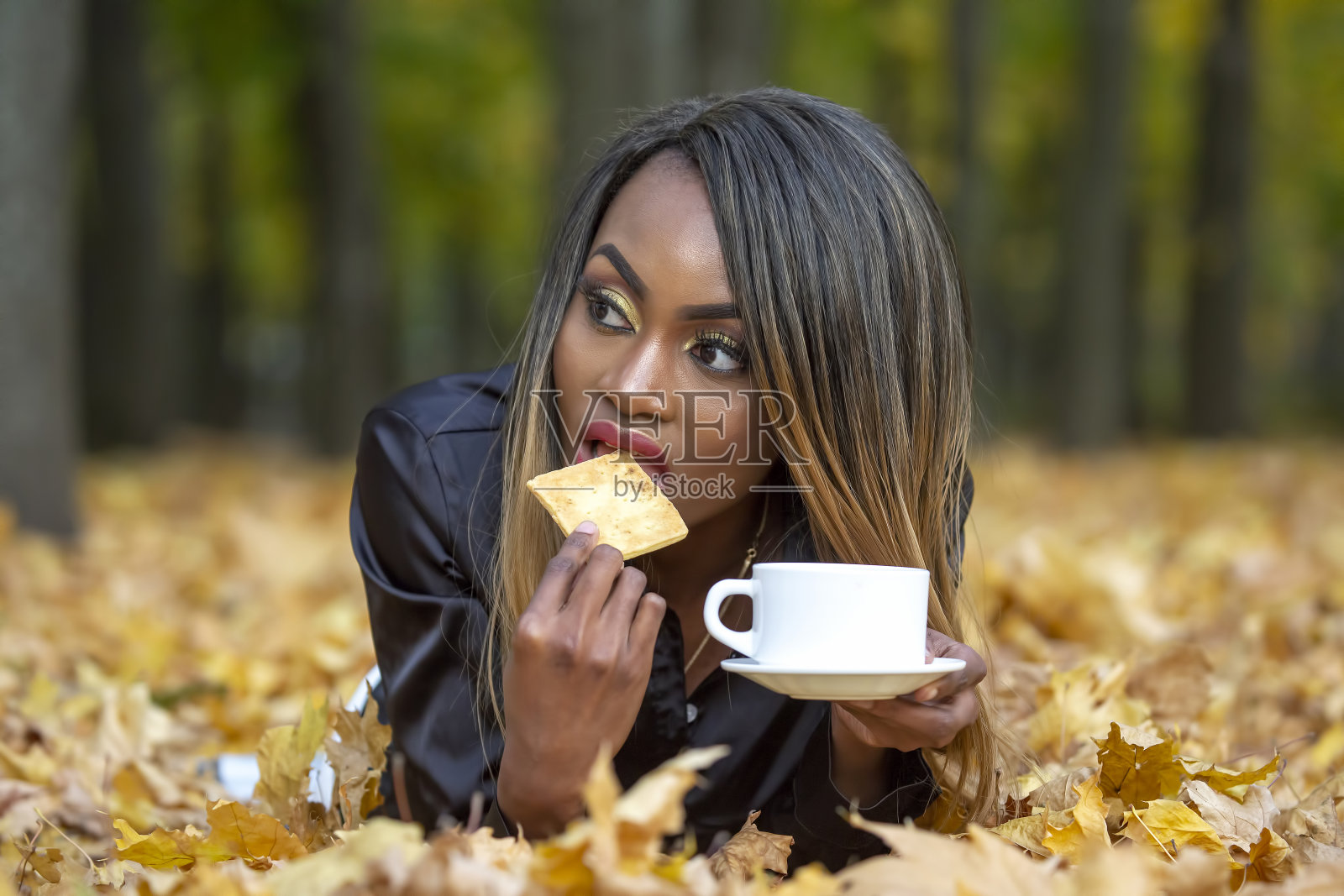 美丽的年轻非洲女孩吃饼干和喝咖啡从白色杯子在秋天的树叶背景在公园照片摄影图片