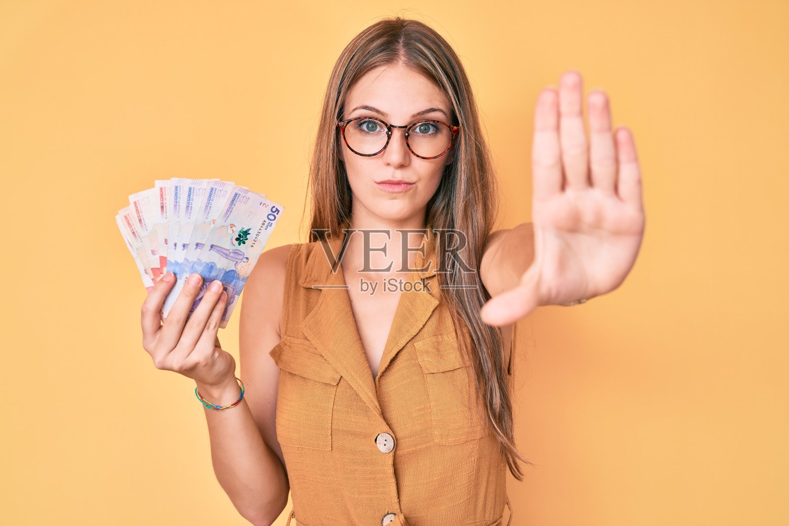 年轻的金发女孩拿着哥伦比亚比索与张开的手做着严肃和自信的表情停止手势,防御手势照片摄影图片