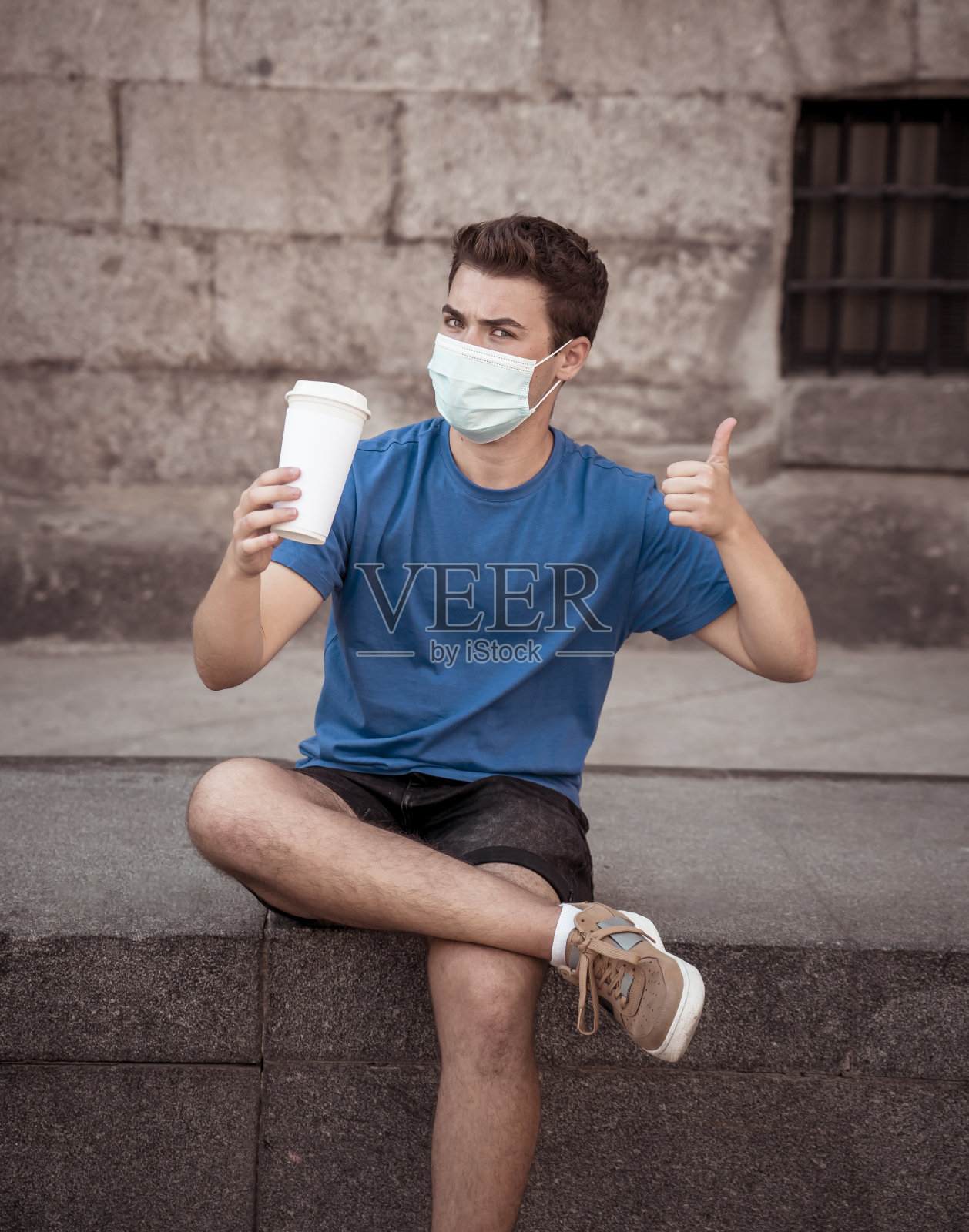 COVID-19暴发。年轻男子戴着外科口罩走在城市街道上,喝着咖啡。冠状病毒、新常态和强制使用口罩照片摄影图片