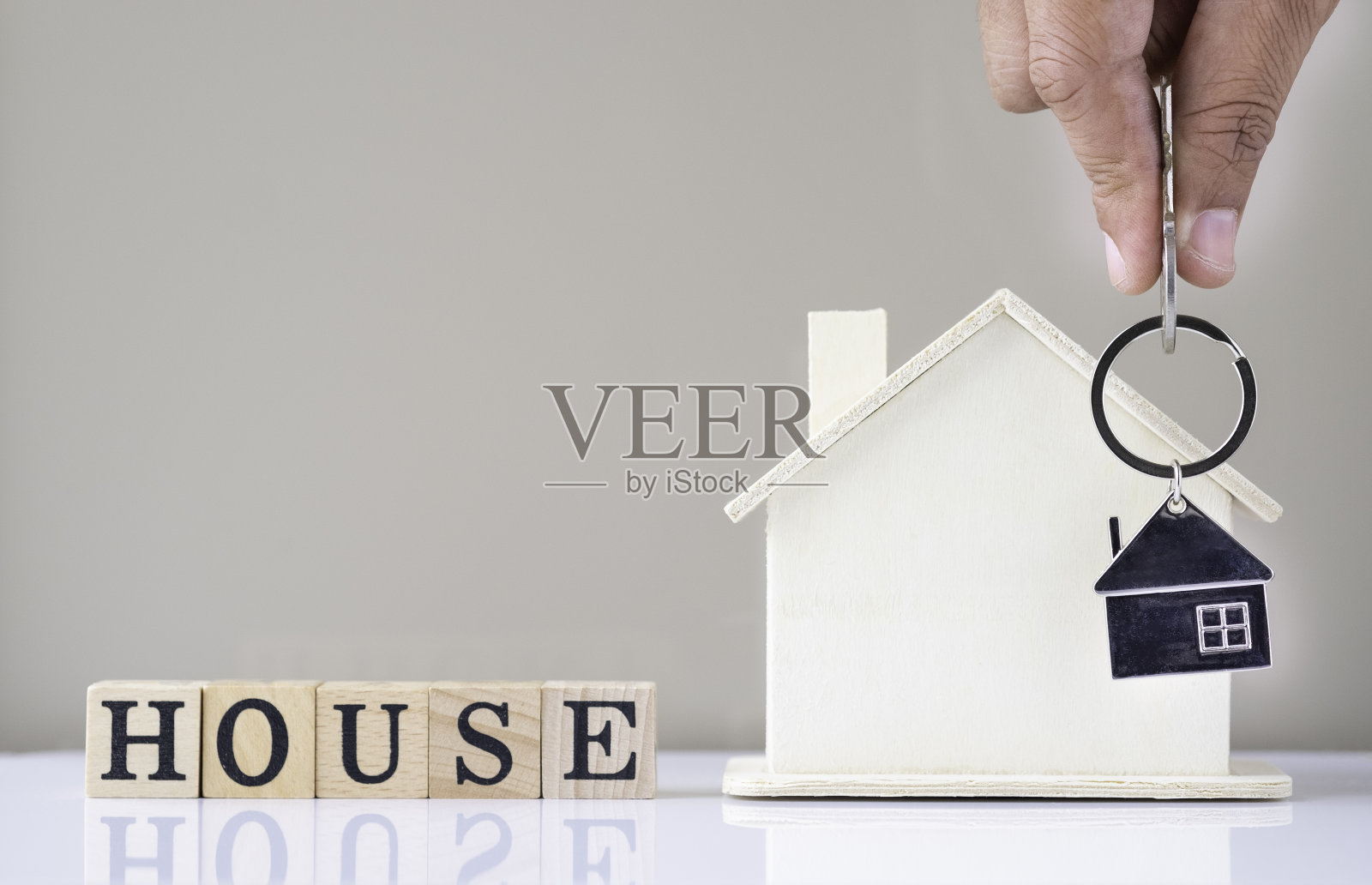 手握银钥匙房子的特写与房子形状的钥匙链和家庭模型在白色的桌子背景。房地产的概念是买一个有拷贝空间的新家。照片摄影图片