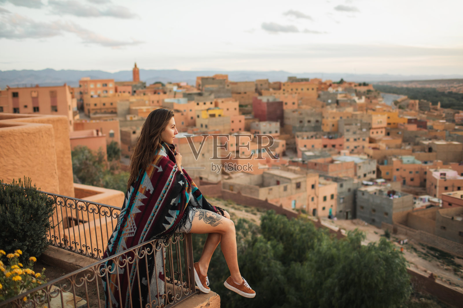 老妇人坐在屋顶露台上鸟瞰阿拉伯古镇布马恩。覆盖着传统的摩洛哥斗篷。嬉皮风格,裸腿纹身。照片摄影图片