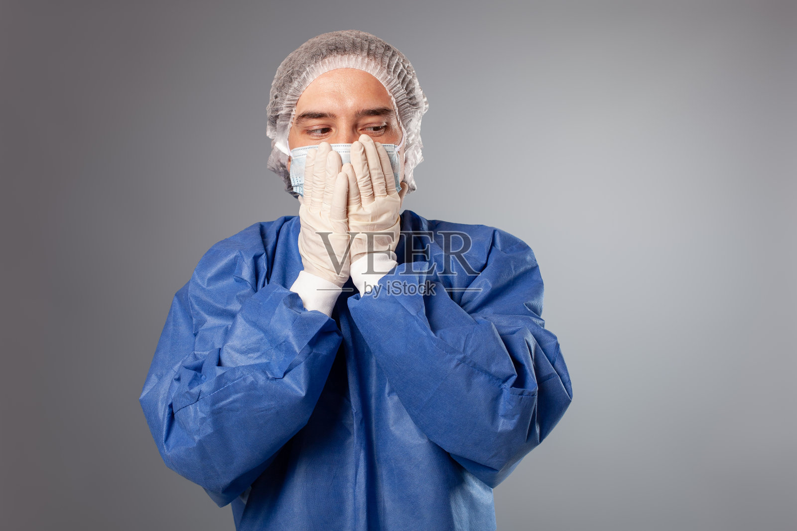 一个英俊的男外科医生是一个医生在一个灰色的背景。一个穿着长袍,戴着医用面具,戴着帽子的人。空的空间。一个人用手捂着嘴,什么也不想说。照片摄影图片