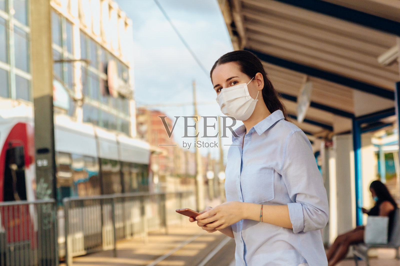 一张年轻女子戴着口罩,拿着电话等着有轨电车到达车站的照片照片摄影图片