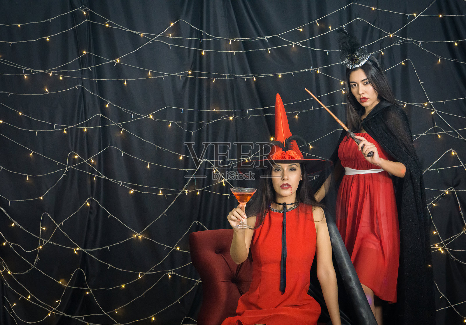 一个穿着红裙子的墨西哥女人拿着鸡尾酒杯坐在沙发上。她的俄罗斯朋友站在后面,手里拿着一根女巫的魔杖。用闪烁的灯光装饰万圣节派对的气氛照片摄影图片