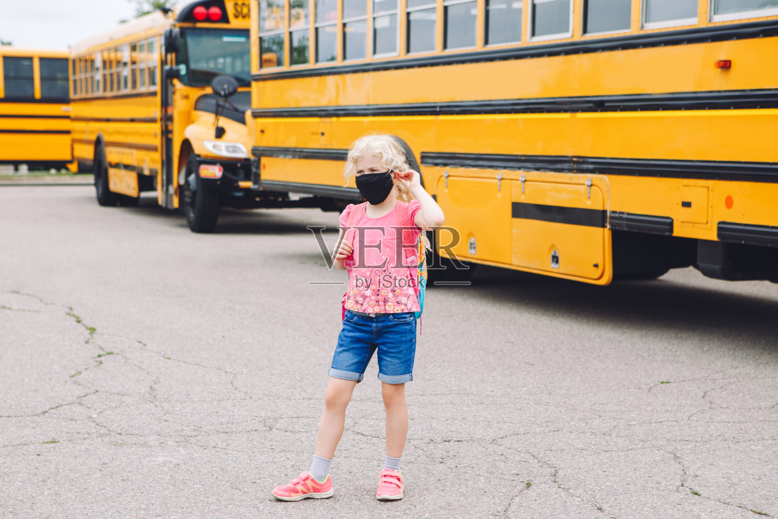 快乐的白人女学生戴着口罩靠近黄色巴士。脸上戴着个人防护装备的孩子。九月入学和返校。冠状病毒感染的新常态。照片摄影图片