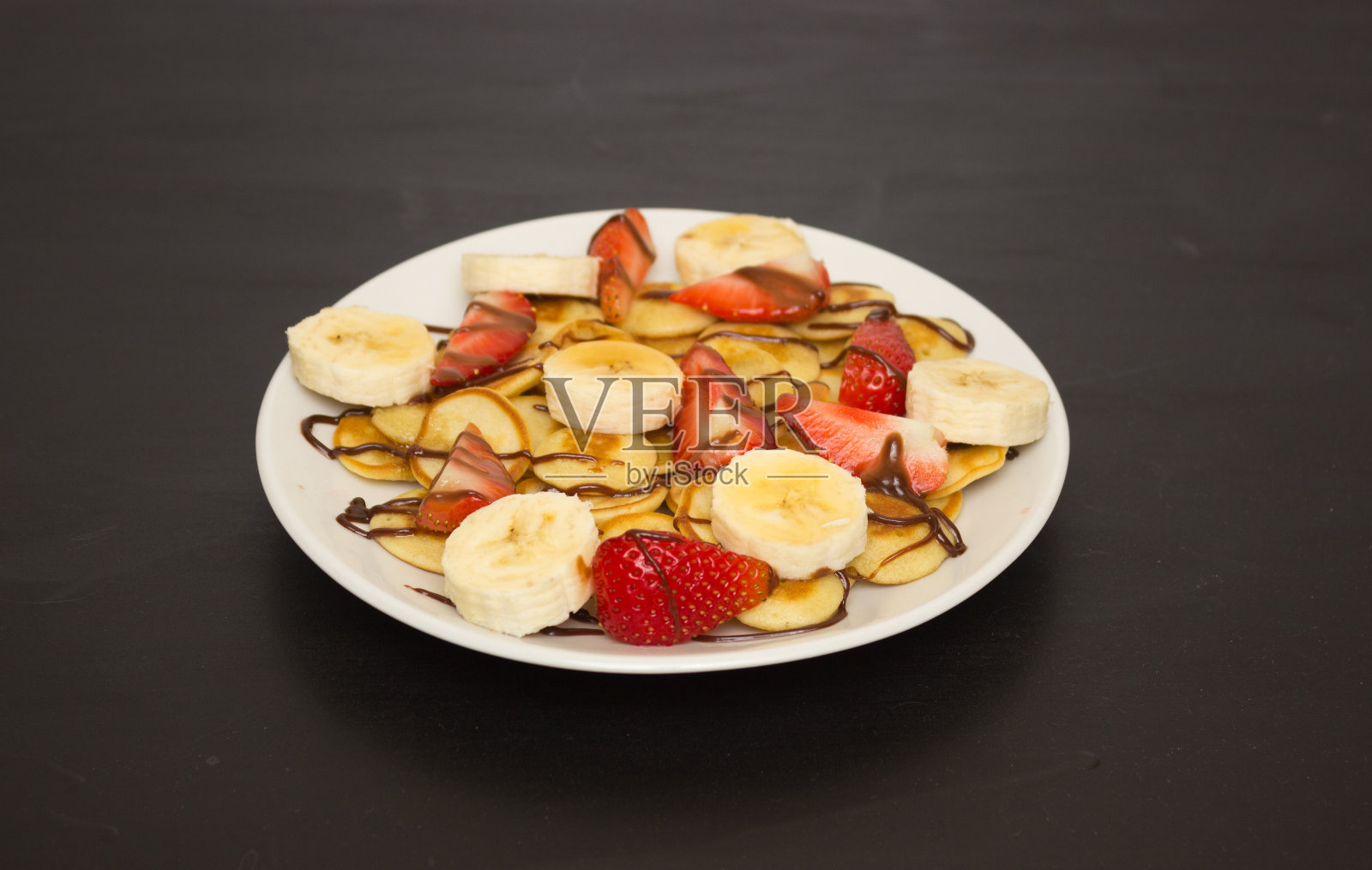 黑色背景的白色盘子上的迷你煎饼。香蕉和草莓片。美味健康的早餐。照片摄影图片