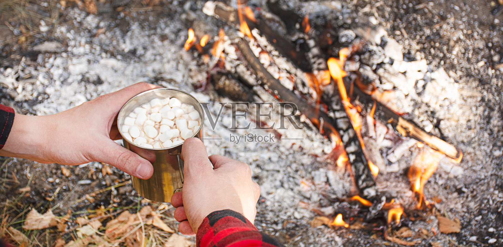 在熊熊燃烧的火焰背景下,男性的手拿着一个可可杯。用金属杯加热可可和棉花糖。秋天的概念。禁令。本空间照片摄影图片