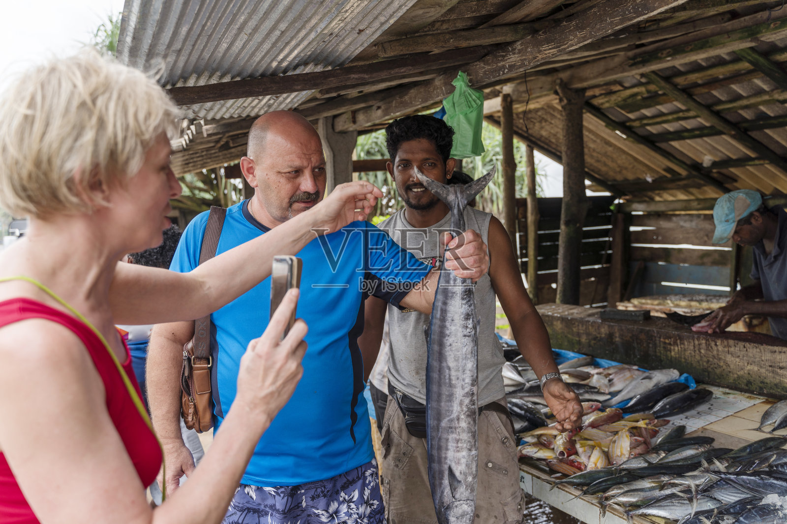 一群欧洲游客,一对夫妇,在斯里兰卡的一个小市场上买新鲜捕到的梭鱼,用智能手机拍照。照片摄影图片