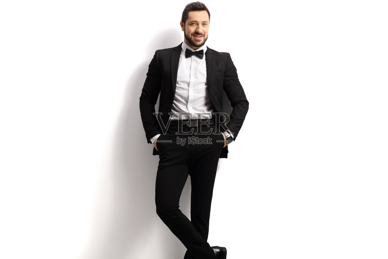 一个帅气的男人,穿着黑色西装,系着领结,倚在一堵白色的墙上照片摄影图片