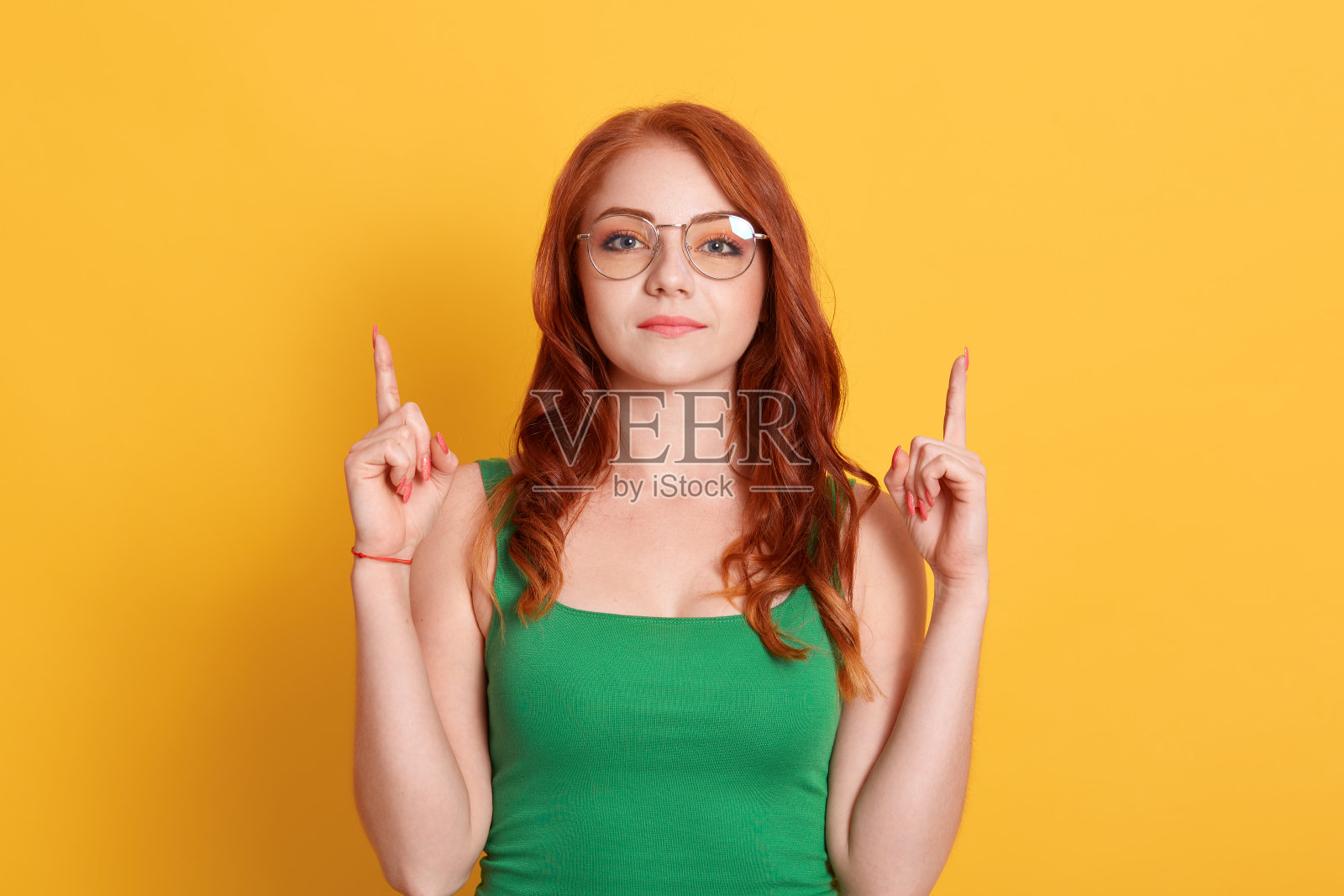 红发严肃的女人穿着绿色衬衫,食指向上,站在黄色的背景,广告版面,漂亮的女孩戴着眼镜。照片摄影图片