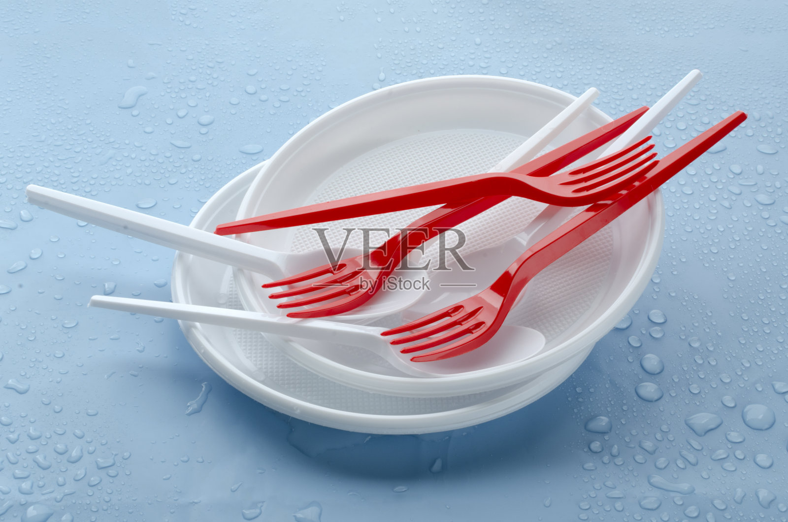 一堆白色塑料盘子和红色,白色的叉子和勺子在潮湿的蓝色桌子上的特写照片摄影图片