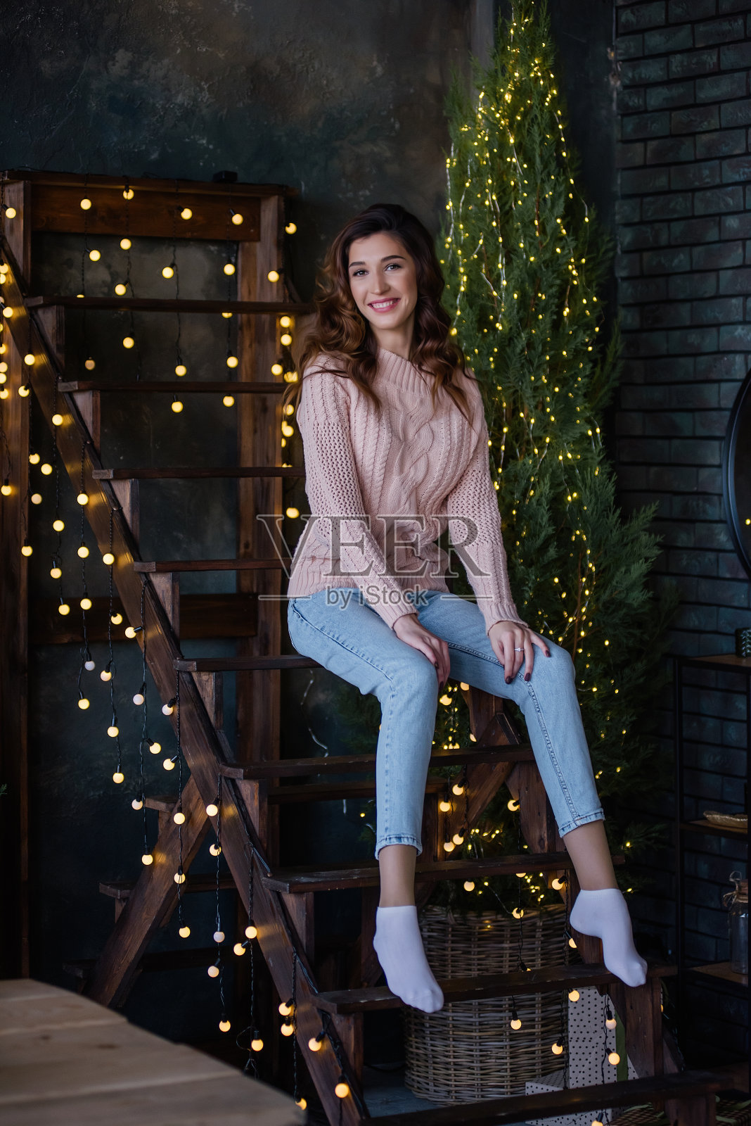 一个女孩,穿着柔软的粉红色针织毛衣,蓝色牛仔裤,留着长长的卷发,坐在木楼梯上,周围是圣诞树和挂着彩灯的花环。年轻女子的特写肖像照片摄影图片