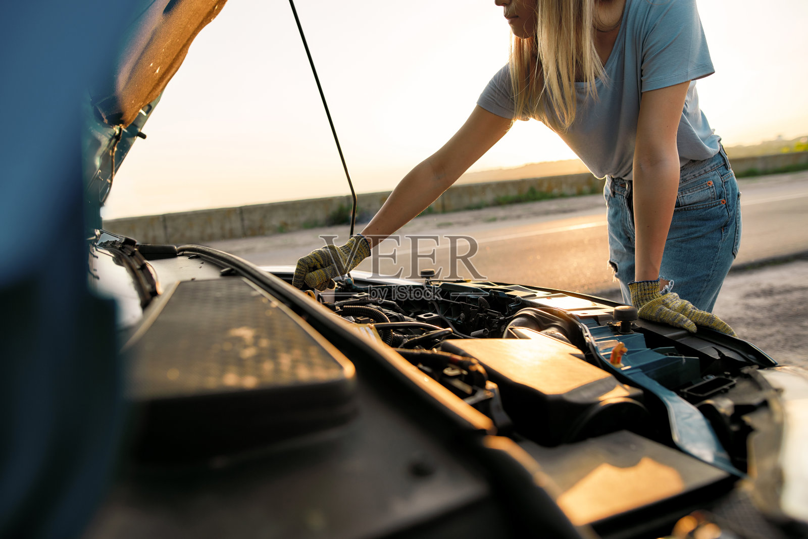 这是一名年轻女子在路边等待救援时,向她的破车引擎盖下面看去,试图自己修理照片摄影图片