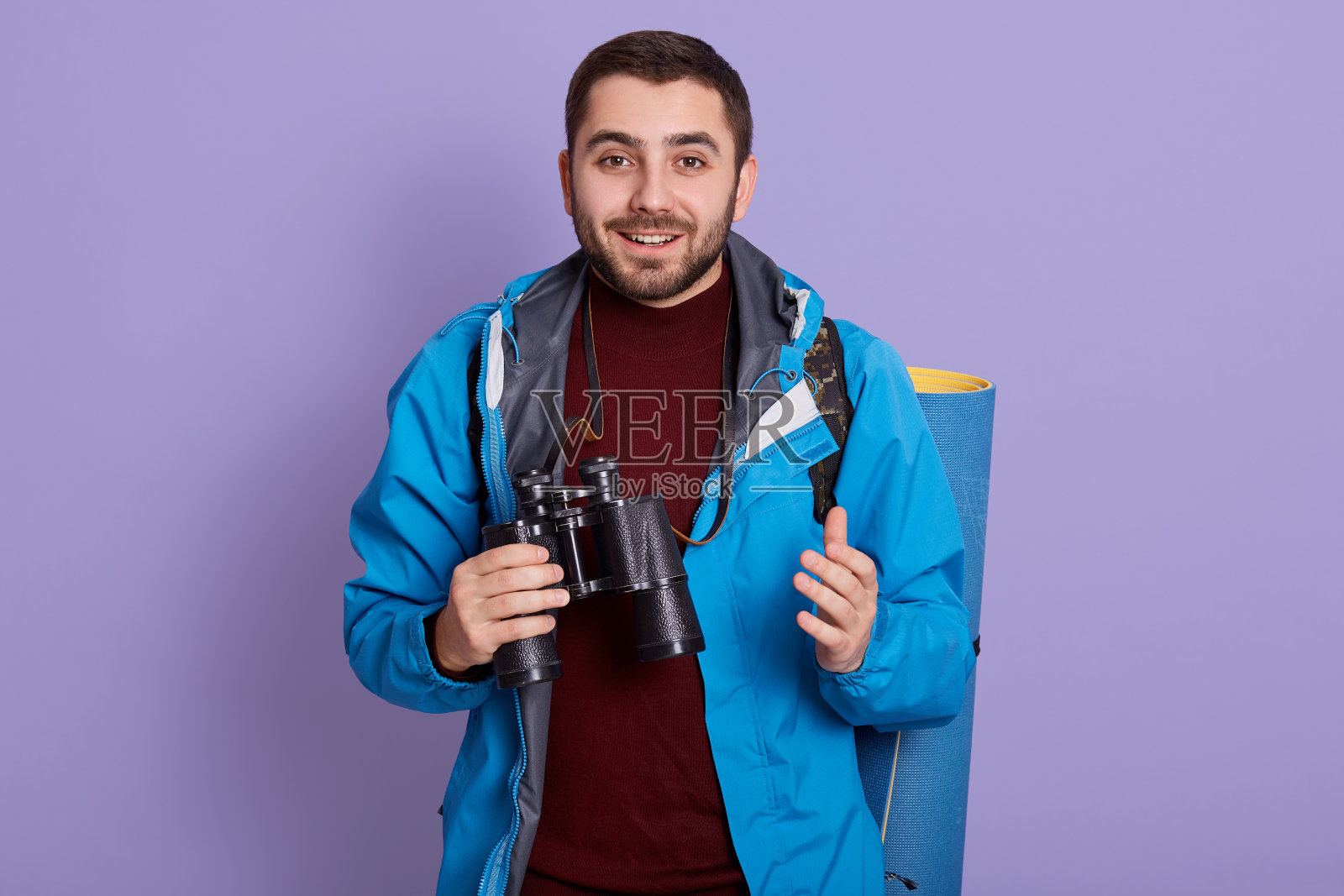 微笑着看相机,手拿双筒望远镜的男游客,穿着夹克和毛衣的大胡子男人,喜欢旅游,喜欢徒步,站在紫丁香墙。照片摄影图片