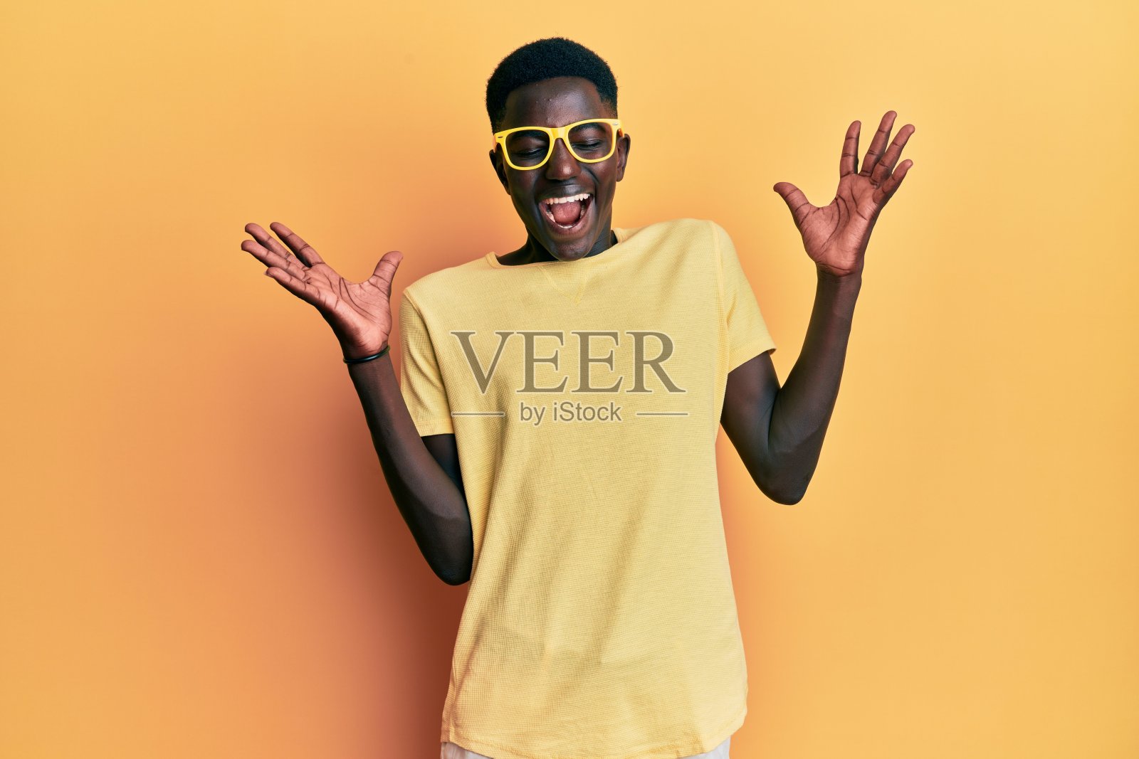 年轻的非洲裔美国人穿着休闲的衣服,戴着眼镜,高举双臂,闭上眼睛,兴奋地尖叫着,为成功而疯狂地庆祝。赢家的概念照片摄影图片