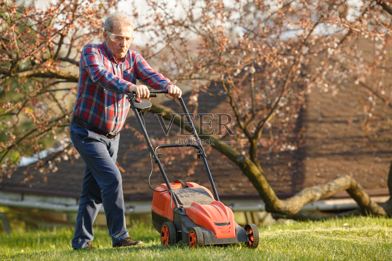 男人拿着电动割草机,割草。修剪花园的园丁。晴天,郊区,村庄。照片摄影图片