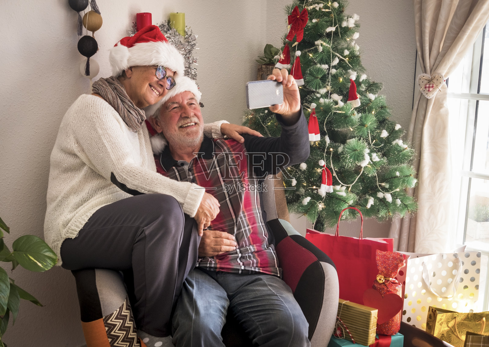 一对戴着圣诞老人帽的老人微笑着坐在同一张扶手椅上,用智能手机自拍。礼物和圣诞树的背景-活跃的社会和科技退休人士照片摄影图片