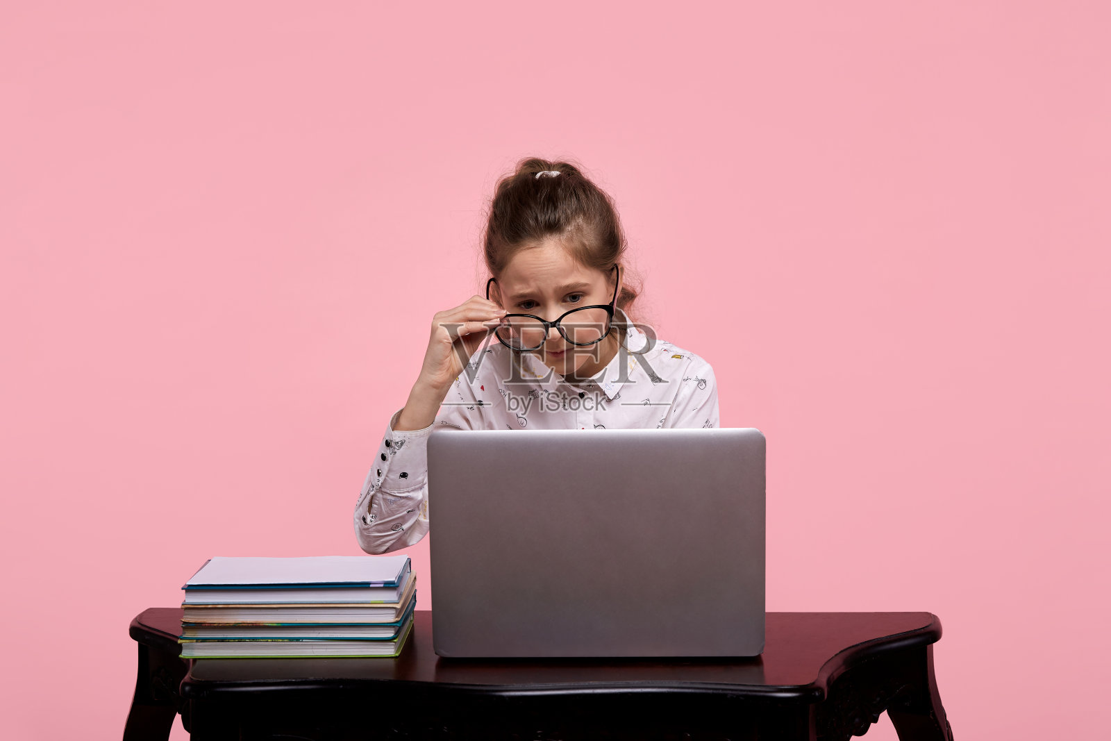 桌子上有一台笔记本电脑和一些书。一个戴着眼镜的漂亮女孩坐着照片摄影图片