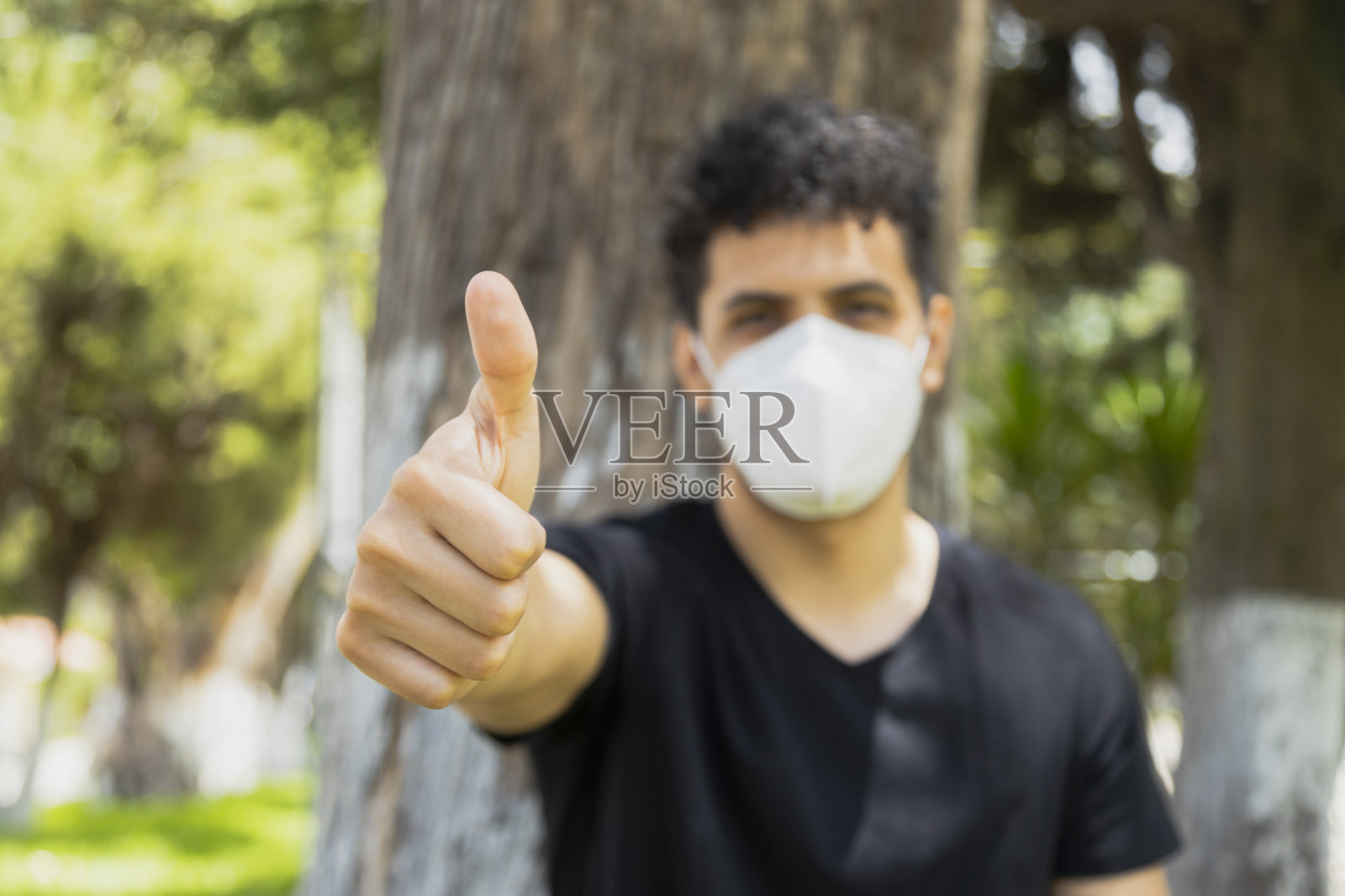 一名西班牙裔青年戴一次性口罩,竖起大拇指,在阳光明媚的日子坐在公园的一棵树下,采取预防措施避免感染任何病毒,社交距离照片摄影图片