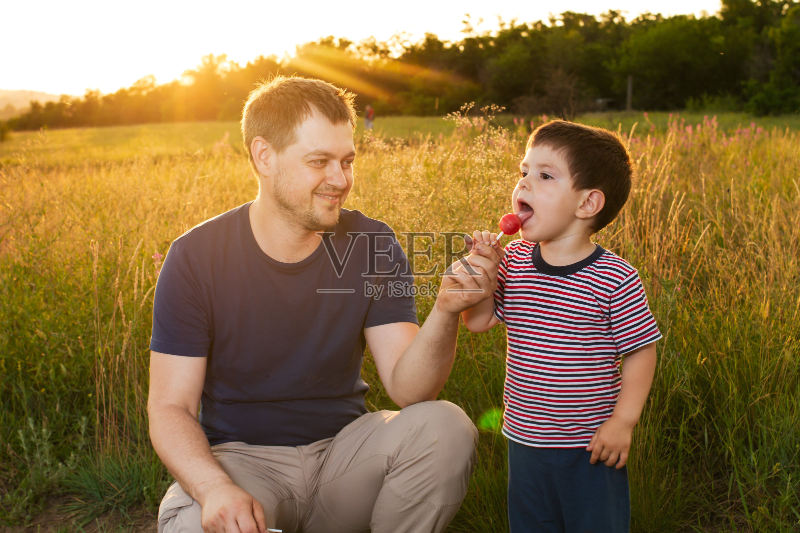爸爸和儿子在傍晚日落时走在黄色的夏日田野上,吃着棒棒糖。照片摄影图片