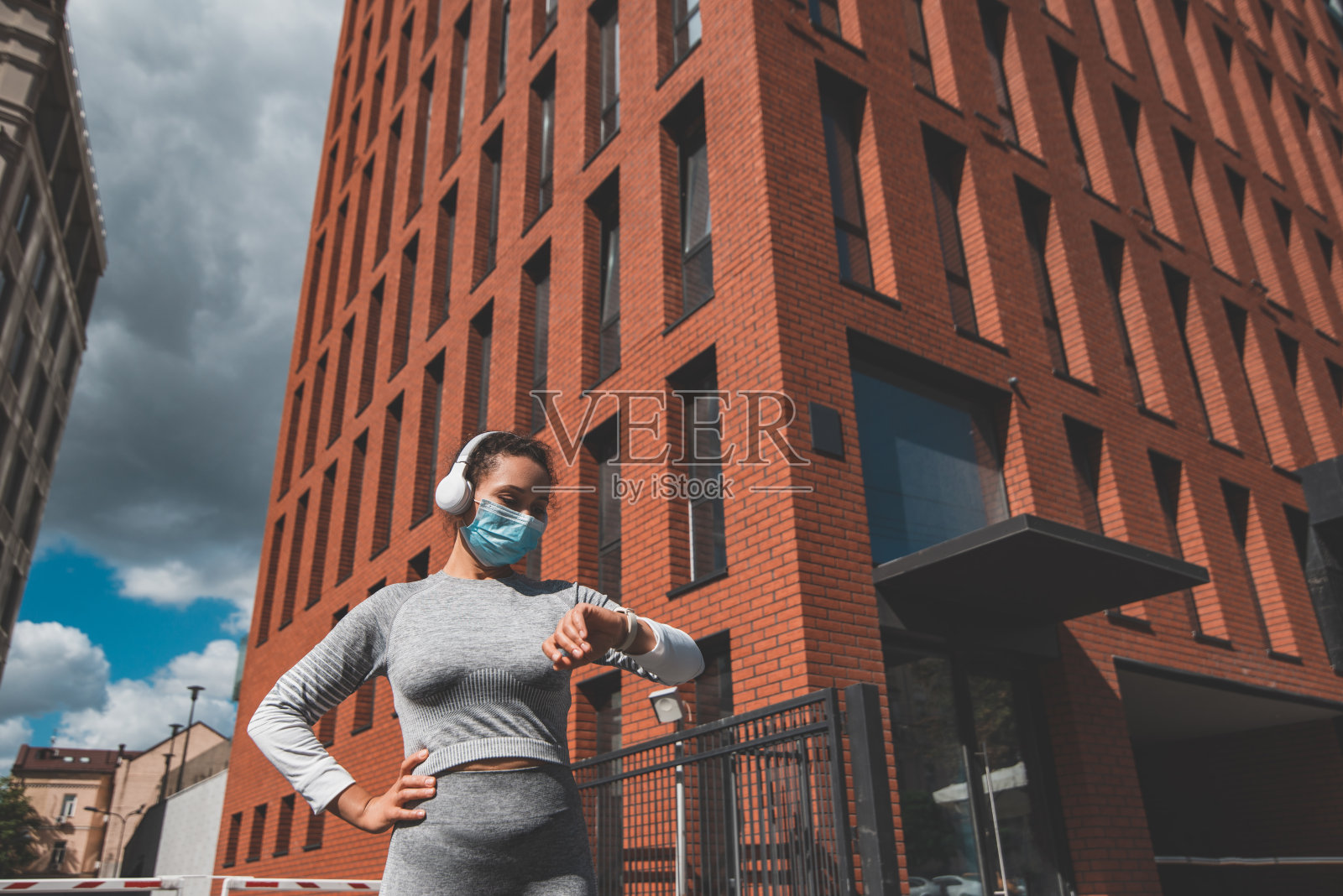 戴着医用口罩戴着耳机的女孩站在一座红砖建筑前照片摄影图片
