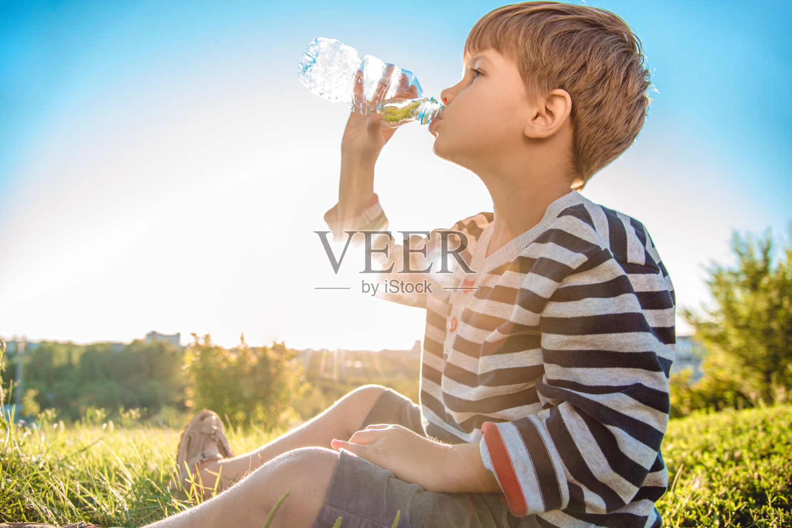 夏日日落时分,可爱的小男孩坐在草地上用瓶子喝水。孩子在热天解渴照片摄影图片