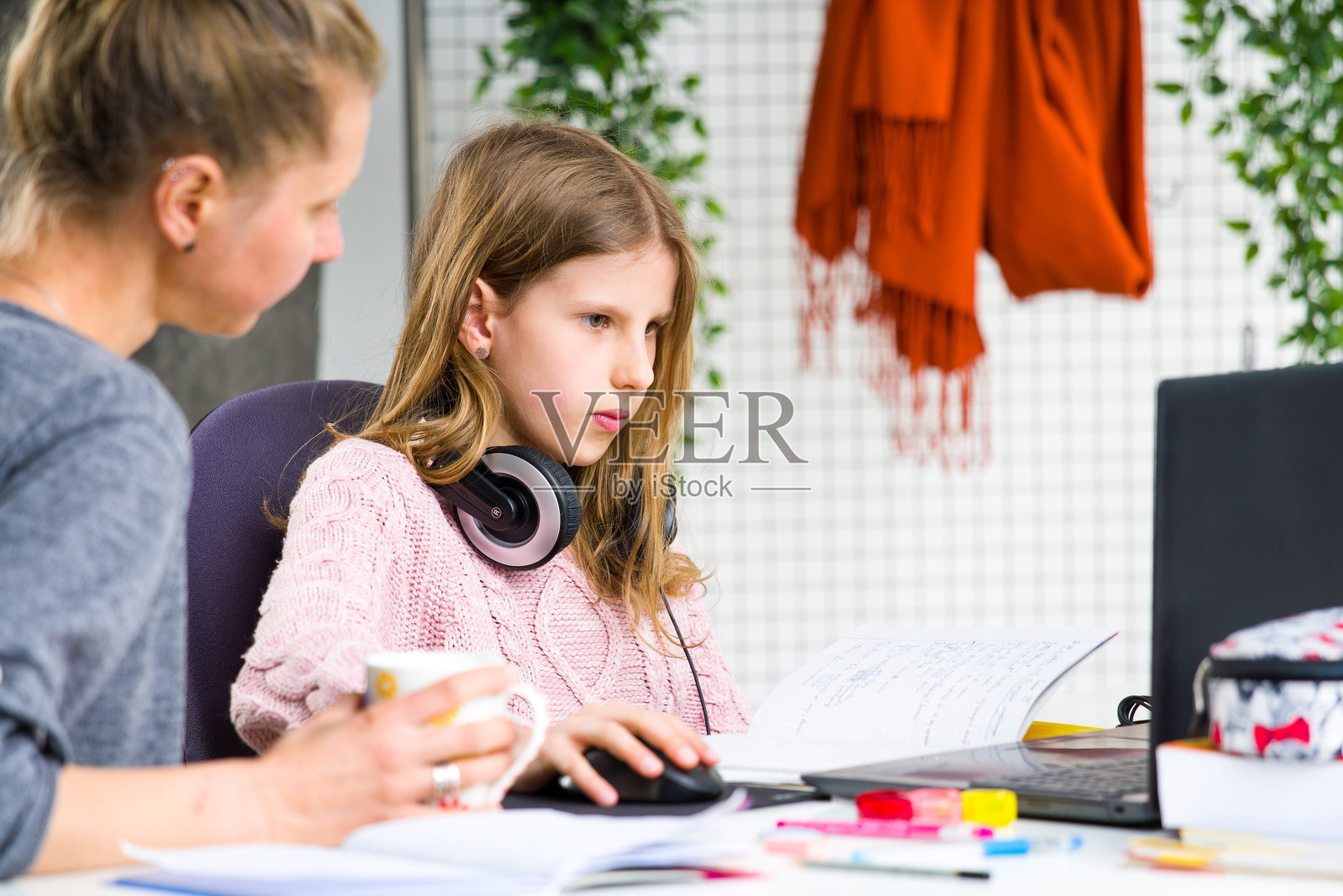 年轻,金发女孩在做她的功课时,粉红色的毛衣是困惑。她妈妈在帮助她。在家隔离期间在家上学。照片摄影图片