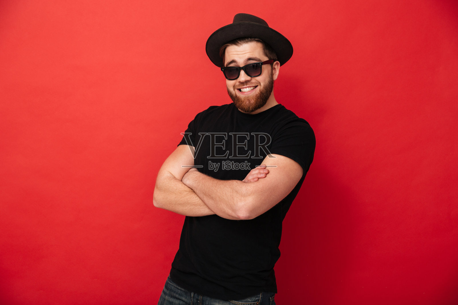 这是一幅不可抗拒的30多岁时尚男士的肖像,他戴着帽子和太阳镜,双手交叉,在镜头前微笑,在红色背景下孤立照片摄影图片