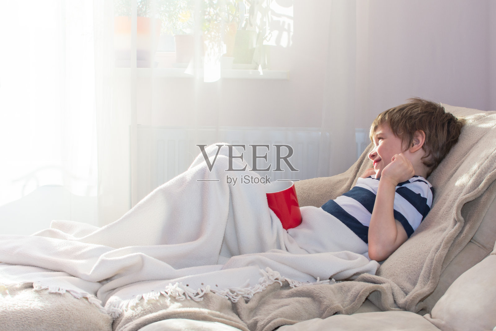 一个阳光明媚的早晨,靠近窗户的地方,男孩躺在床上,盖着一条毯子。照片摄影图片