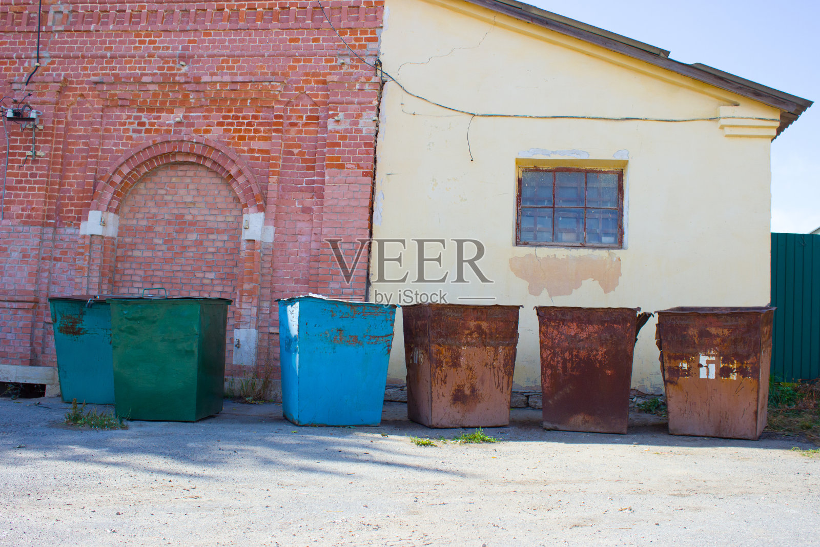 在一个阳光明媚的夏日里,彩色的旧空垃圾桶映衬着一座砖房和一座用黄色灰泥砌成的建筑。照片摄影图片